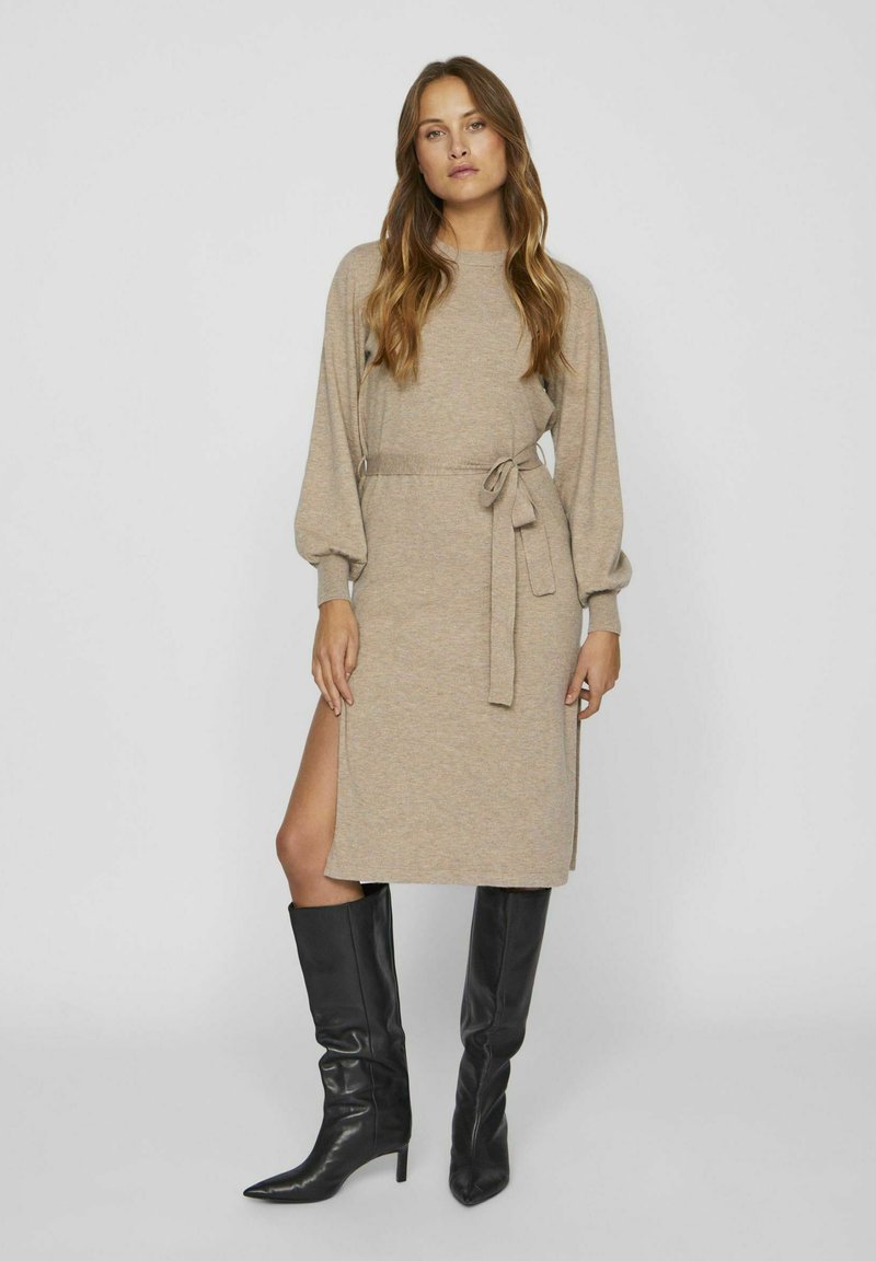 Vestito a maglia beige con maniche lunghe, cintura in vita e spacco laterale; abbinato a stivali neri in pelle al ginocchio. Tessuto liscio e design aderente.