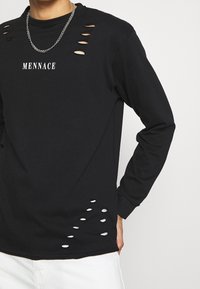 Černé tričko s dlouhým rukávem s výřezovými detaily kolem výstřihu a bočního lemu, s potištěným logem "MENNACE" v bílé barvě.