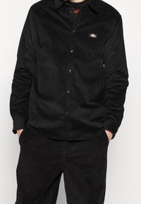 Camicia in velluto a coste nero con chiusura frontale a bottoni, caratterizzata da una piccola toppa logo sul petto sinistro. Maniche lunghe e collo classico.