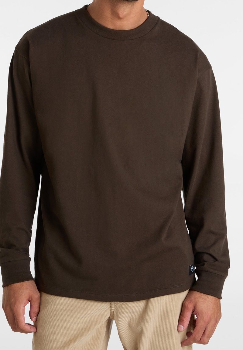 Mies, ktorý má na sebe jednoduchú tmavohnedú košeľu s dlhým rukávom a béžové kordové nohavice, stojí na bielom pozadí.