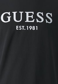 Sort t-shirt med stort hvidt tekst der står "GUESS" og "EST. 1981." Stoffet ser glat ud med en afslappet pasform.
