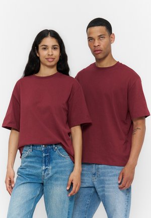 To personer står side om side iført matchende bordeauxfarvede t-shirts og lyseblå jeans mod en ensfarvet hvid baggrund.