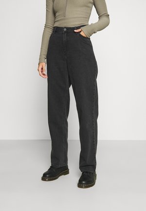 Vrouw draagt losse zwarte jeans met hoge taille, olijfgroene geribbelde top met lange mouwen en zwarte veter schoenen, staat met één hand in haar zak.