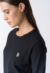 Maglione nero a maglia con scollatura rotonda, caratterizzato da un piccolo logo dorato ricamato sul petto. Texture morbida e design aderente.