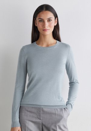 Kobieta o długich ciemnych włosach ubrana w jasnoniebieski sweter z okrągłym dekoltem i szare spodnie stoi na białym tle, z jedną ręką w kieszeni.