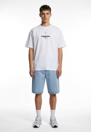 Jeune homme debout, portant un t-shirt blanc "Urban Classics", un short en jean bleu clair, des chaussettes blanches, des baskets blanches et un collier avec un pendentif.