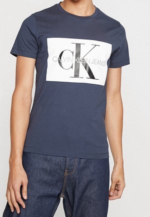 Osoba nosząca granatowy t-shirt Calvin Klein Jeans z dużą białą naszywką z logo oraz ciemnoniebieskie jeansy.
