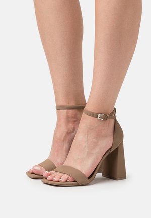 Sandales à talons hauts beiges avec un orteil carré, une large sangle à l'avant, une sangle de cheville et un talon carré. Texture lisse avec une finition mate.