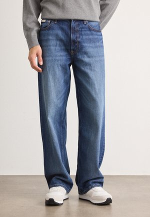 Mann trägt locker sitzende blaue Jeans, grauen Pullover und weiße Sneakers, steht auf einem Betonboden vor weißem Hintergrund.