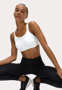Vit sportbh med övre panel av mesh, justerbara axelremmar och krokuppkoppling. Kombinerad med högmidjade svarta leggings och sneakers.