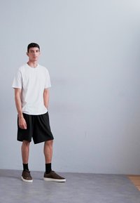 White short-sleeve t-shirt, black shorts, and brown sneakers with textured surface. Standing on gray flooring against a light gray wall.