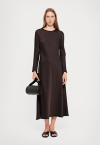 Langermet, gulvlengde kjole laget av glatt, mørk brun stoff. Modellen holder en svart, sylinderformet håndveske og har på seg flate, svarte sandaler.