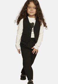 Bambino che indossa un gilet nero con bottoni sopra una camicia a maniche lunghe bianca con grafica, abbinato a pantaloni neri e scarpe nere.