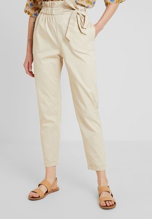 Frau trägt beige Papiertaschen-Hochbundhose mit gebundenem Gürtel und hellbraunen Sandalen mit offenem Zehenbereich, steht vor einem einfarbigen Hintergrund.