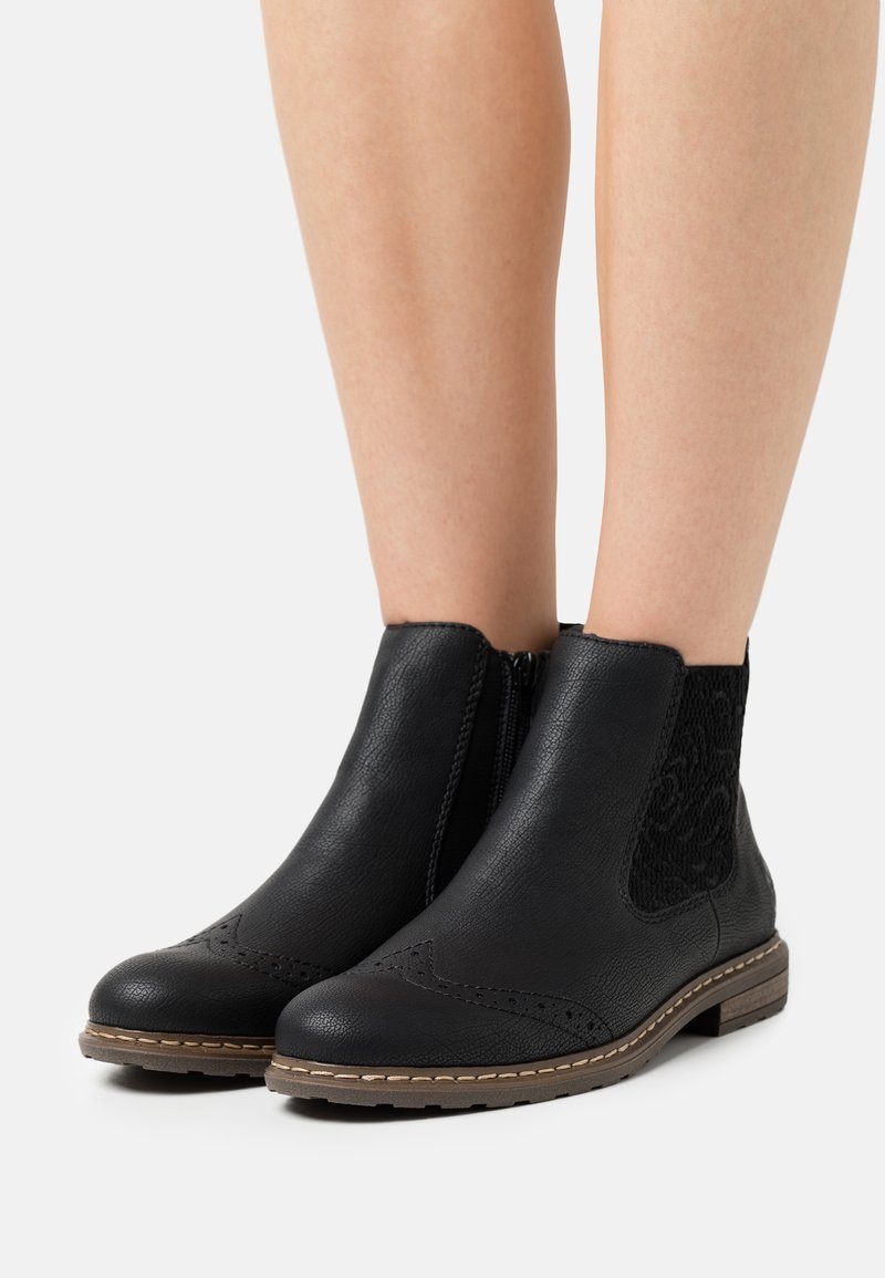 Bottines noires avec fermeture éclair latérale, cuir texturé, coutures décoratives, panneau jacquard élastique et semelle basse marron portées sur jambes nues.