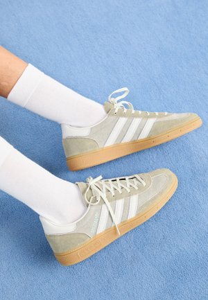 Pieds portant des chaussettes blanches et des baskets en daim gris clair avec des bandes blanches et des semelles en gomme sur fond de tapis bleu.