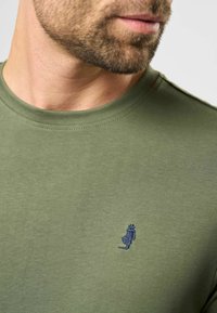 Olivinės spalvos marškiniai su trumpomis rankovėmis, pagaminti iš minkštos medvilnės. Kairėje krūtinės pusėje yra mažas tamsiai mėlynas siuvinėjimas. Apvalus kaklas.