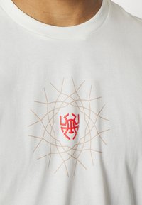 Valkoinen puuvillapaita, jossa on keskellä punainen logo, ja sen ympärillä on geometrisista linjoista muotoiltu pyöreä hämähäkkiverkko design vaaleanbeigen värisenä.