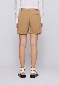 Person som bär beige uppvikta shorts, vit långärmad skjorta och vita sandaler med svarta sulor, stående mot en enkel bakgrund.
