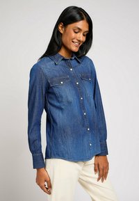 Camicia di denim con un gradiente di blu chiaro a blu scuro, chiusura frontale con bottoni, due tasche sul petto e colletto classico con maniche lunghe.
