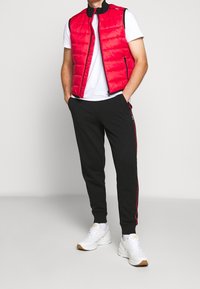 Gilet imbottito rosso con colletto alto, indossato sopra una maglietta bianca. Pantaloni atletici neri con strisce laterali rosse. Sneakers bianche completano il look.