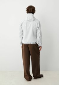 Ljusgrå jacka med huva, som har en mjuk yta och elastiska muddar. Bärs med bruna, löst sittande byxor. Minimalistisk design.