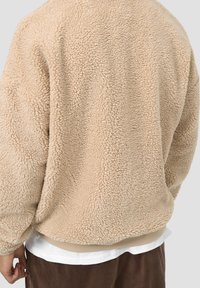 Pullover in pile beige con una superficie morbida e strutturata. Spalle scese e orlo a coste. Indossato sopra una camicia bianca. Design con schiena arrotondata.