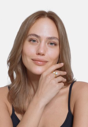 Anello d'oro con due perle bianche, indossato su una mano, con una texture liscia e lucida. La pelle è leggermente luminosa su uno sfondo semplice.
