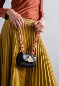 Geantă mică neagră matlasată cu logo auriu, accentuată de o bretea de lanț multicolor. Poartă lângă o fustă plisată muștar.
