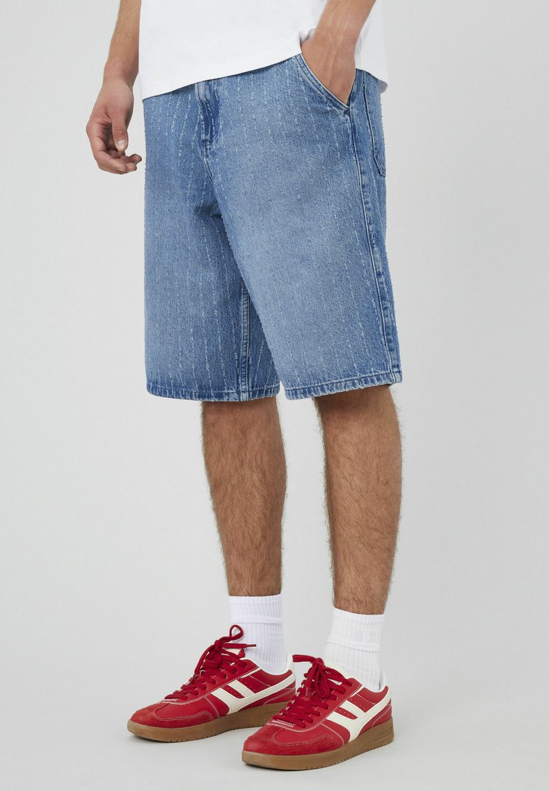 Shorts en denim bleu avec une coupe décontractée, présentant un léger délavage et des détails de texture. Associés à des chaussures de sport rouges et blanches et des chaussettes blanches.