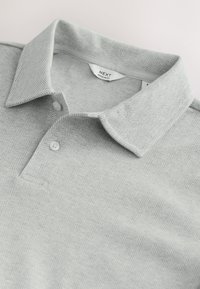 Polo gris clair avec un motif chevrons subtil, deux boutons et une étiquette "NEXT Regular Fit" au col.