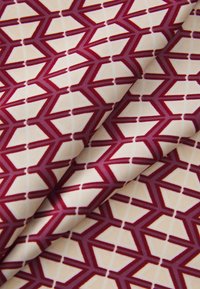Tissu imprimé présentant un design géométrique avec des formes bordeaux entrelacées sur un fond crème. Texture lisse, matériau tissé.