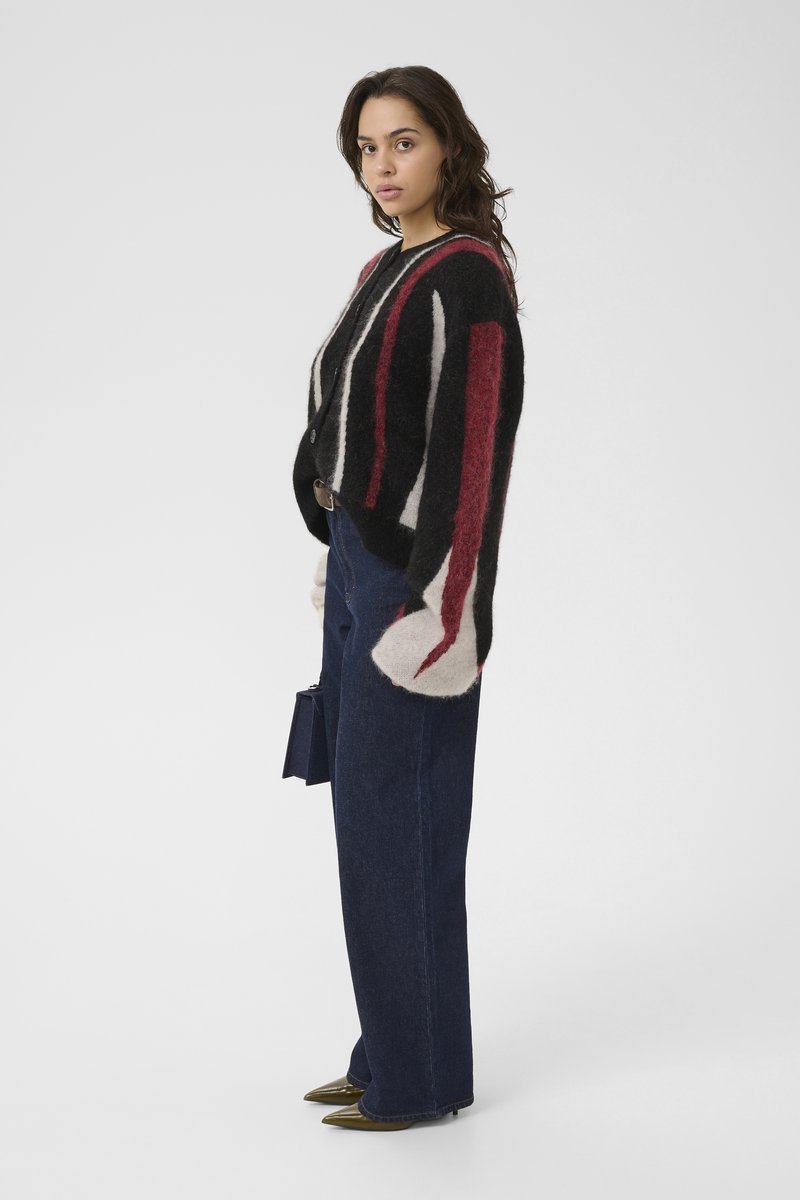 Donna in profilo che indossa un cardigan morbido a strisce nere, bianche e rosse, jeans scuri a gamba larga, scarpe a punta e tiene una piccola borsa.