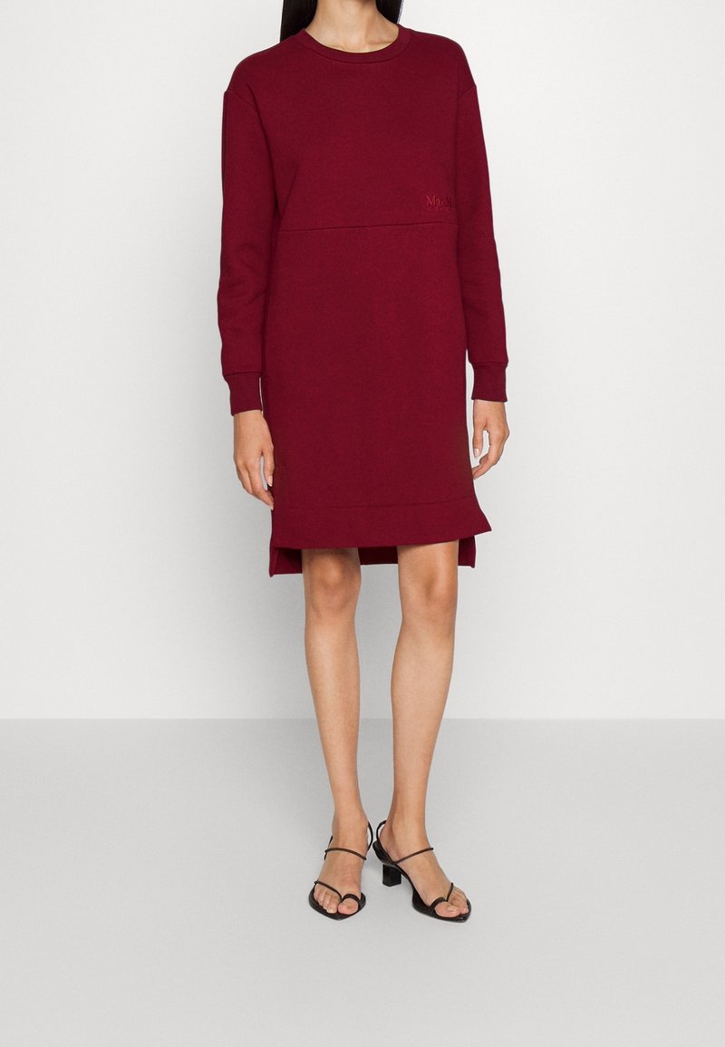 Femme portant une robe bordeaux à manches longues, longueur genoux, avec un ourlet asymétrique et des sandales noires à talons avec des brides.