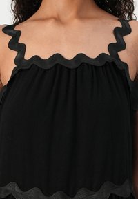 Μαύρο τοπ με ρέουσες τιράντες και χαλαρή γραμμή. Το ύφασμα φαίνεται απαλό και ελαφρύ, με λεία υφή και χωρίς ορατά μοτίβα.