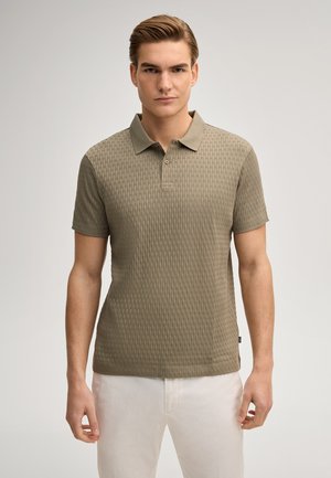 Giovane uomo con polo a maniche corte verde oliva testurizzato e pantaloni bianchi, in piedi davanti a uno sfondo chiaro e semplice.