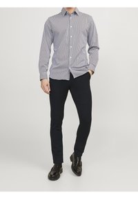 Muž oblečený v černých slim fit kalhotách, černých formálních botách a dlouhém košili s knoflíky s tenkými vertikálními pruhy, stojící s jednou rukou v kapse.