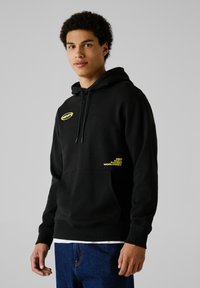 Sudadera negra con bolsillo delantero, capucha con cordón y detalles de logo amarillo. Fabricada en una suave tela texturizada. Combinada con jeans azules.
