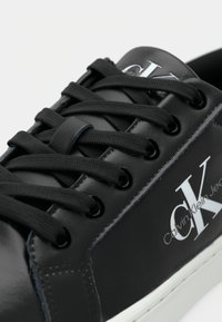 Detailný záběr na čiernu koženú tenisku Calvin Klein s čiernymi šnúrkami a bielou podrážkou, logo viditeľné na boku a jazyku.
