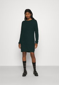 Vestido de malha riscado em verde escuro, com mangas longas, decote redondo e comprimento até à coxa. Usado com meias de padrão xadrez e loafers pretos.