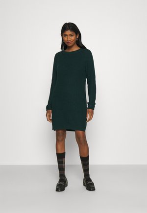 Robe tricotée côtelée, vert foncé, avec des manches longues, un col rond et une longueur mi-thigh. Portée avec des chaussettes à carreaux et des mocassins noirs.