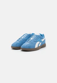 Sapatos atléticos em camurça azul com detalhes brancos, apresentando um design de atacadores e uma sola de borracha castanha. Textura suave e bico arredondado.