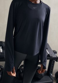 Černý sportovní top s dlouhým rukávem z hladké látky, s kulatým výstřihem a logem Nike. Nošený s černými legínami, ukazující přiléhavý střih.