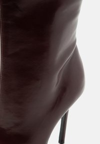 Gros plan d'une botte à talon haut en cuir marron foncé montrant une texture lisse et une silhouette courbée sur un fond clair.