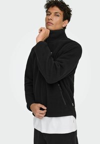 Crna flis jakna s visokim ovratnikom, s patentnim zatvaračima i glatkom unutrašnjom teksturom, nosi se preko bijele majice.