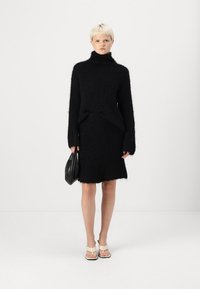 Black knitted sweater dress with a high collar, textured fabric, and long sleeves. Paired with a black handbag and beige sandals.