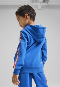 Niebieska bluza z kapturem, z czerwoną i żółtą taśmą z logo biegnącą wzdłuż rękawów. Miękki, teksturowany materiał i luźny krój. Dopasowane niebieskie spodnie.