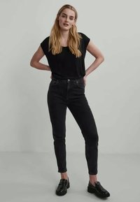 Camiseta preta de manga curta, combinada com jeans pretos de cintura alta. O modelo usa sapatos moccasin pretos, posando contra um fundo cinza claro.