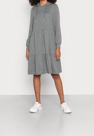 Kobieta ubrana w szarą sukienkę dresową do kolan z długimi rękawami i białe trampki, stojąca na tle jednolitego białego tła.