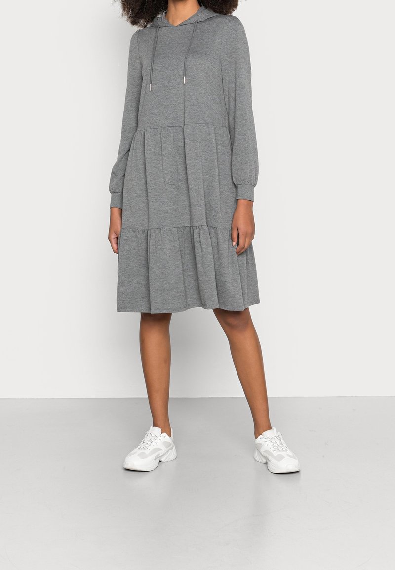 Femme portant une robe à capuche grise mi-longue avec manches longues et baskets blanches, debout devant un fond blanc uni.